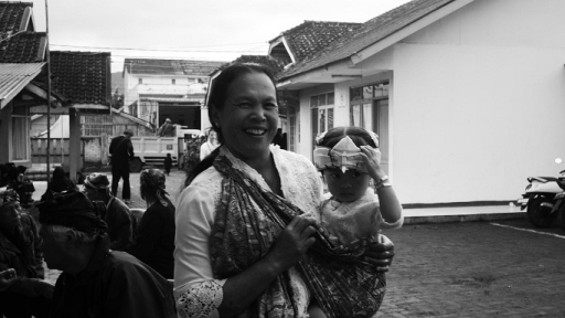 Seorang ibu menggendong anaknya di halaman Pasewakan Waruga Jati, Cicalung bersiap-siap untuk arak-arakan ke sumber mata air. Selasa, 1 Juli 2025. (Foto: Fitri Amanda/BandungBergerak)